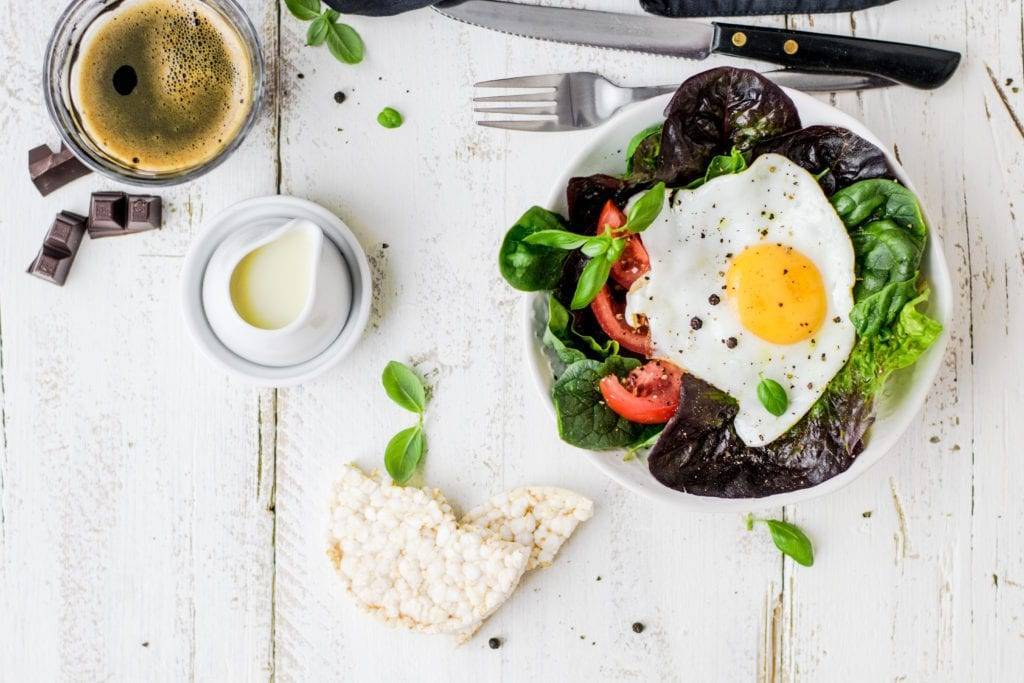 Looking down on table with salada with tomatoes and sunny side up egg with rice cake,cup of coffee cream and fork and knife