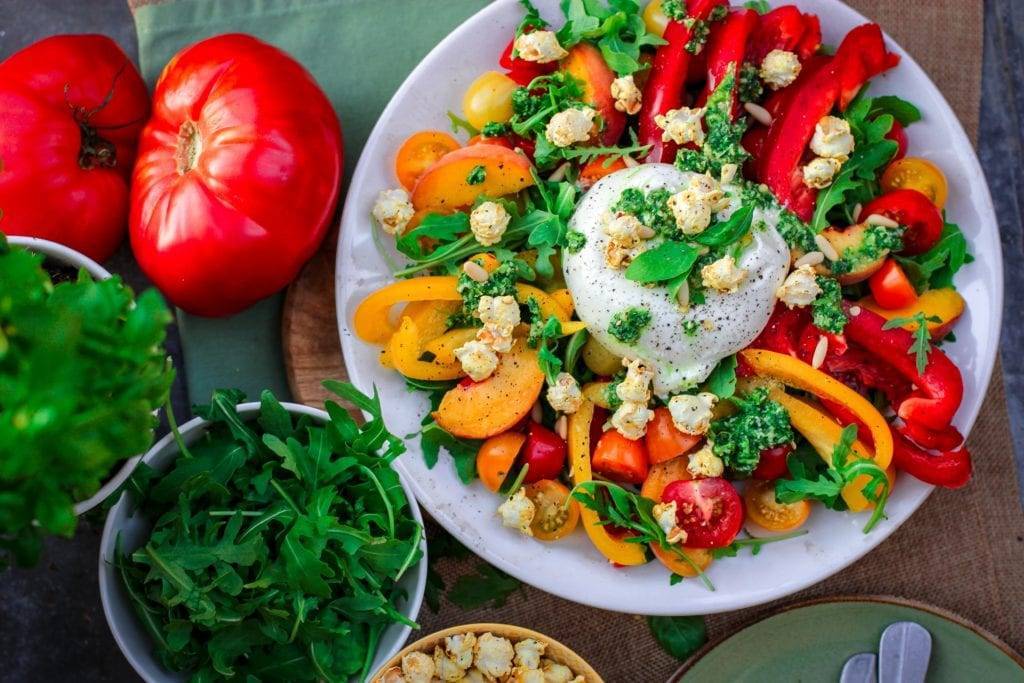 Top view of white bowl filled with sliced orange and red vegetables on table with whole tomatoes and bowls of herbs.