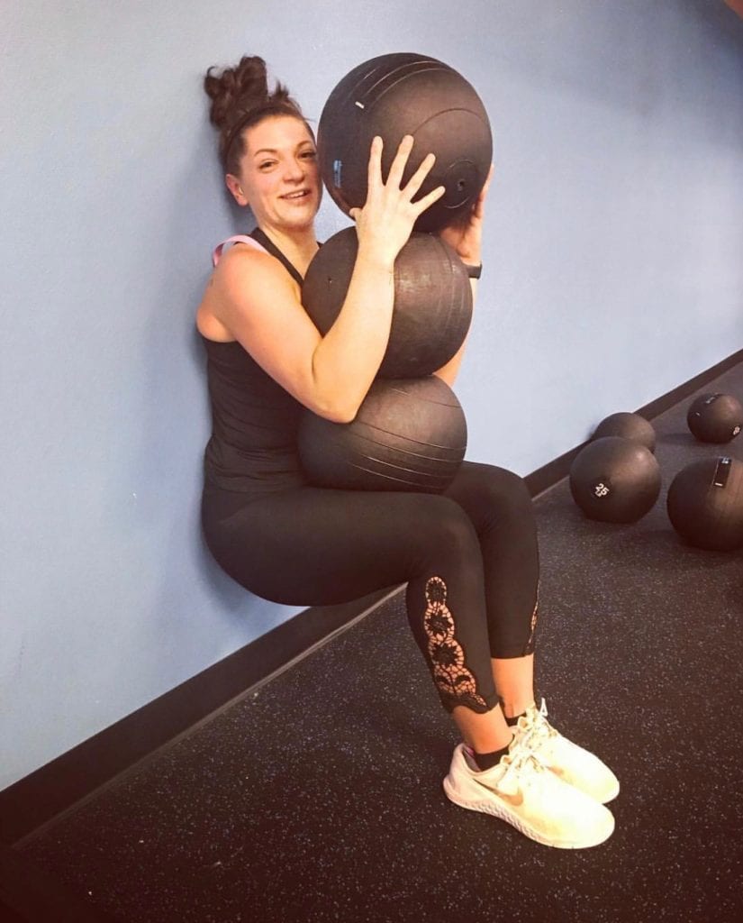 Women with hair in ponytail leaning against a grey wall wearing black tank top and pants with white shoes using 3 medicine balls