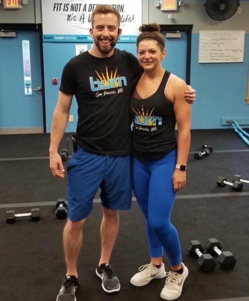 Man with arm around women wearing black Burn Boot Camp shirts standing in gym with black floor and blue walls