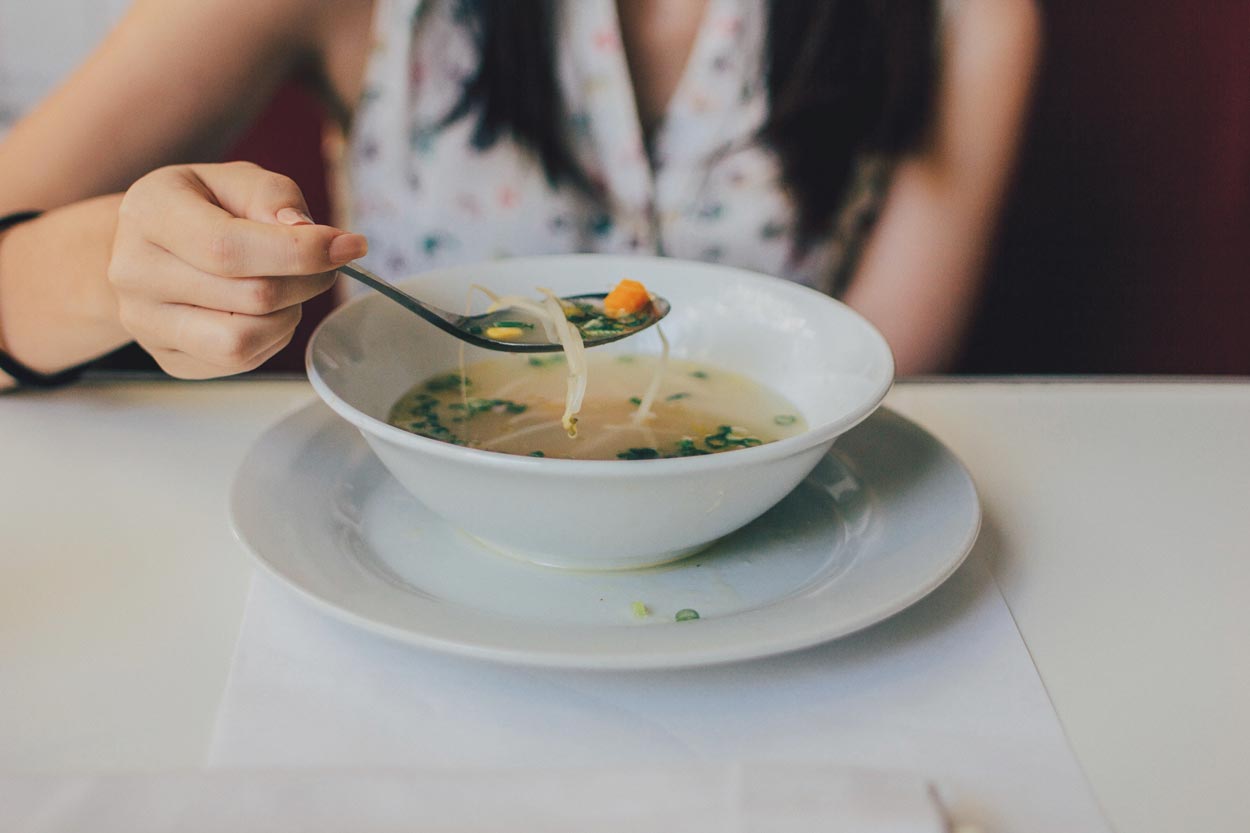 White glass bowl filled with soup on white glass plate on white table with women holding a spoon full of soup over bowl.