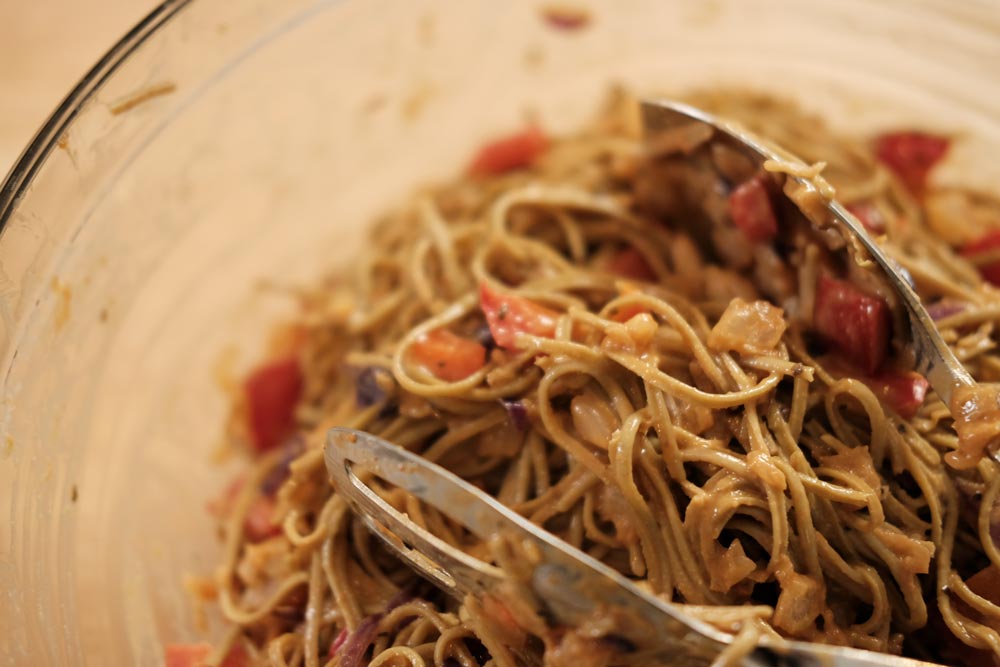 Clear glass bowl of pasta mixed with sauce and sliced tomatoes with mixing tongues.