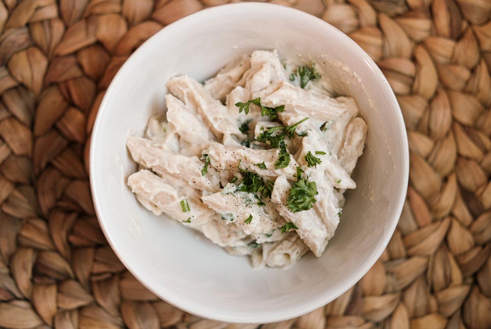 White glass bowl of pasta with creamy white sauce and broccoli on top of table.