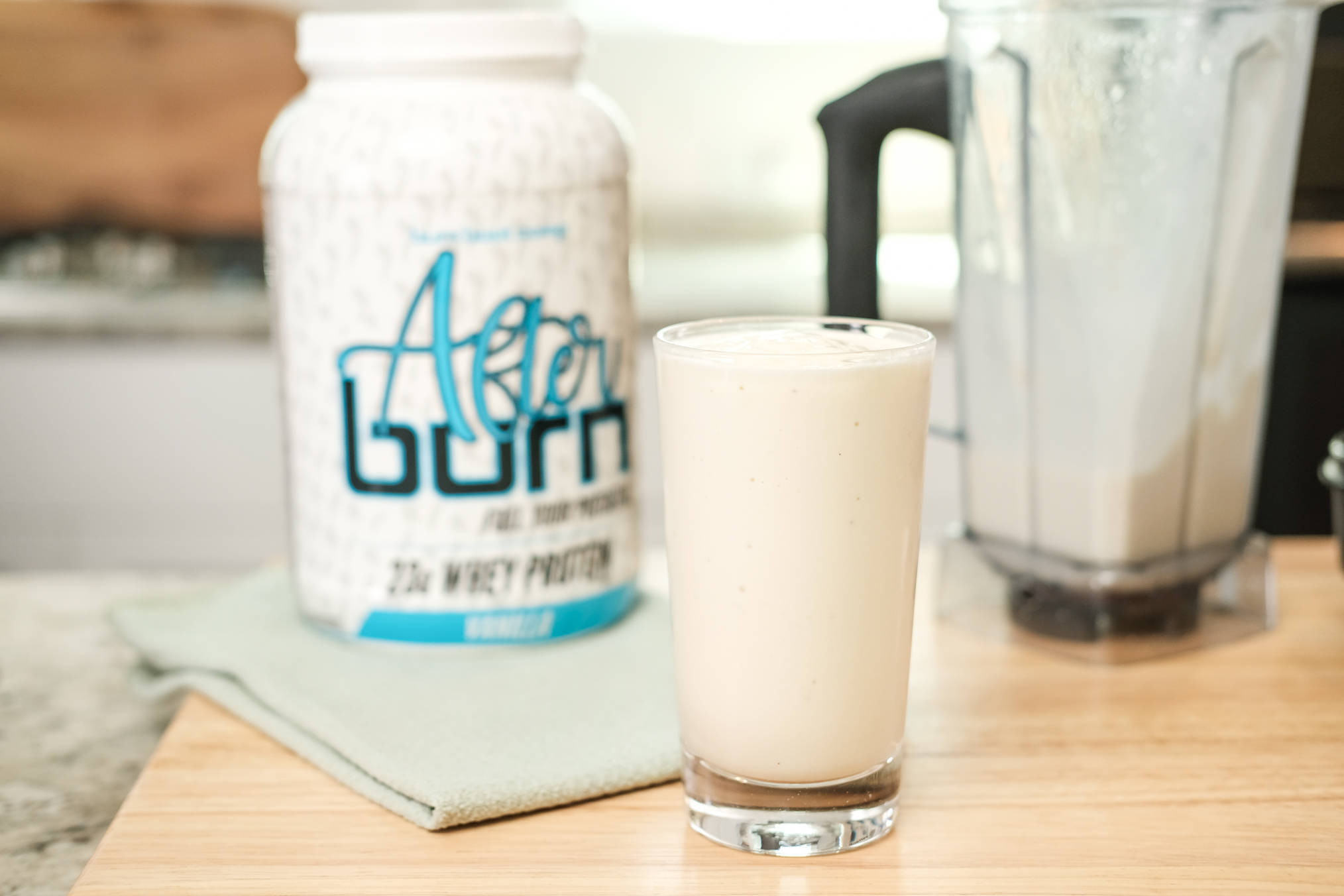 Clear glass filled with pina colada protein smoothie on cutting board on countertop next to white vanilla Afternoon bottle and empty blender.