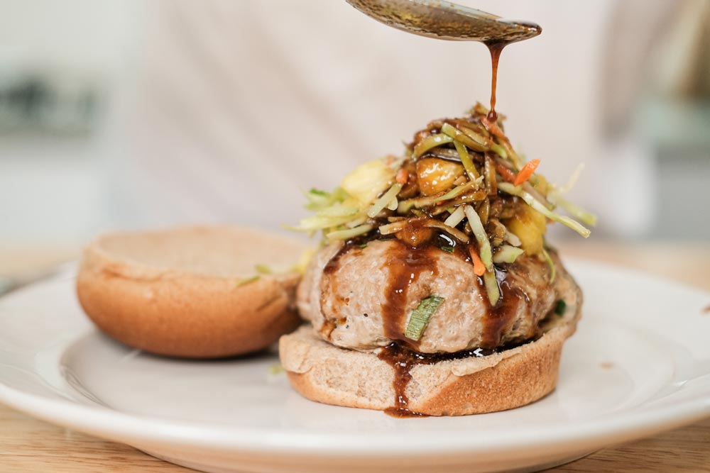 Spoon drizzling dark sauce over turkey burgers with asian salad on top sitting on white glass plate on table