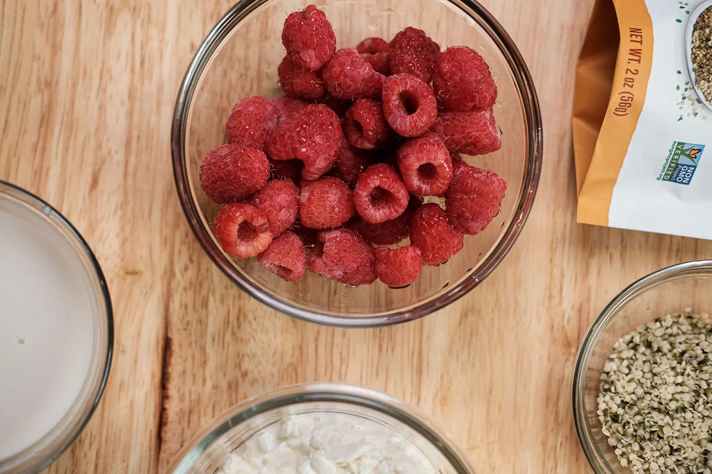 Clear glass bowl filled with fresh raspberries next to three other clear glass bowls filled with oats and milk and cream cheese.