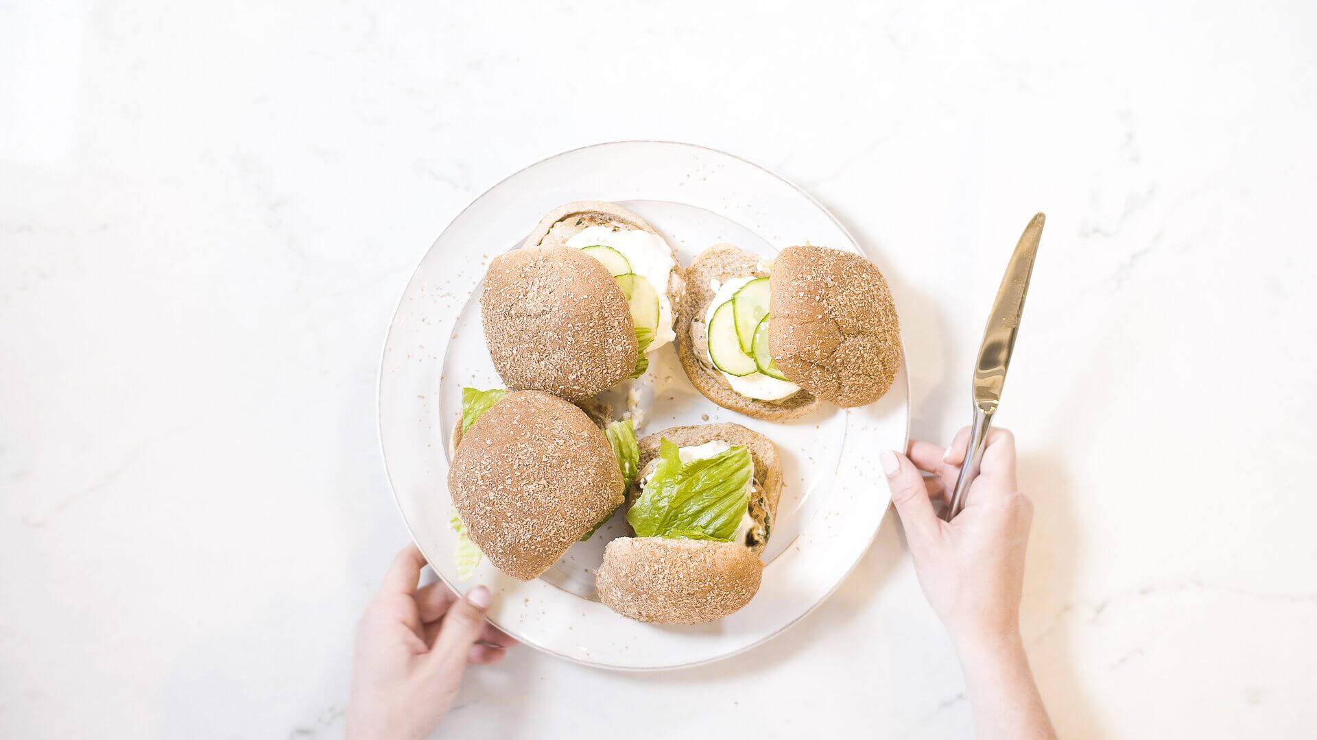 Women's hands with knife in right hand taking a white glass plate with 4 multi-grain sandwiches with cheese and cucumber and lettuce.