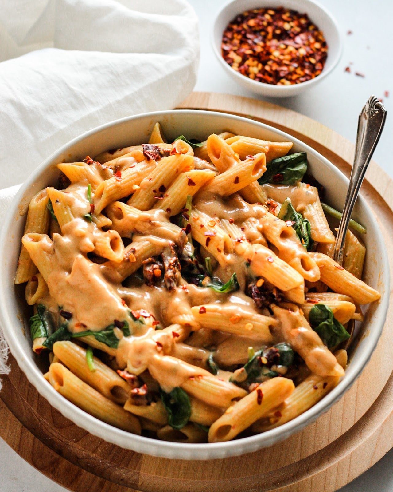 White glass bowl of penne pasta with spicy cream sauce with spinach with silver fork next to bowl of chili flakes.