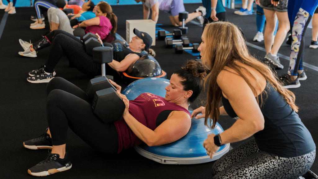 A Burn Boot Camp Trainer assisting a Member in glute bridges as part of our lower body dumbbell workouts.